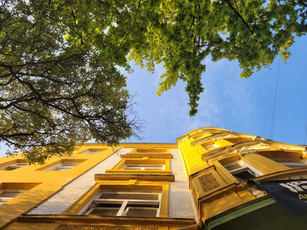Eine gelb-weiße Häuserfassage eines Gründerzeithauses, fotografiert von unten nach oben. Blauer Himmel, Baumblätter.