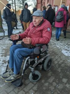 Ein älterer Herr, rote Jacke, graue Schiefermütze, sitzt im Rollstuhl, der auf einem Bürgersteig stet. Im Hintergrund weitere Demonstrierende.