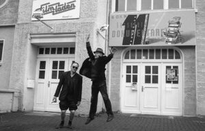 Sw-Bild: Vor einer Hausfassade auf der ein Schild mit der Beschriftung"Filmladen" steht, steht ein kleiner Mann mit Sonnenbrille und kurzen Hosen. Ein weiterer Mann neben ihm springt mit erhobenen Armen in die Luft.
