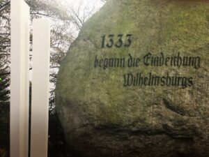 Links sieht man 2 Stelen des Flutdenkmals und über 3/4 des Bildes Ausschnitte des Steins mit der Inschrift: 1333 begann die Eindeichung Wilhelmsburgs