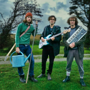 Drei junge Männer stehen mit Gießkanne, E-Gitarre und Synthesizer auf einer Wiese