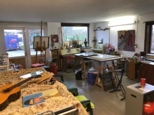 Atelierraum mit ganz viel Kunstwerken. Links ein Gestell mit einer beigen Decke. Darauf eine Gitarre und ein Portrait.