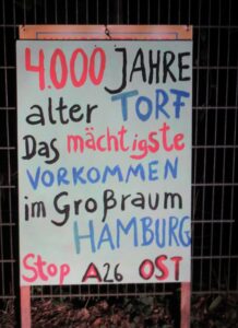 Stellschild mit der bunten Aufschrift: 4000 Jahre alter Torf Das mächtigste Vorkommen im Großraum Hamburg. Stop A26 Ost