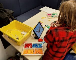 Auf einem Tisch legen in verschiedenen Sortierschalen Legoteile und ein Tablet mit einer Bauanleitung. Ein Kind setzt Teile zusammen.