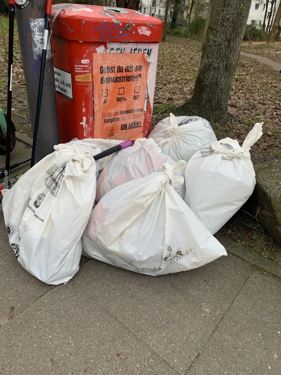 Vor einem roten Stadtmülleimer liegen mehrere weiße gefüllte Mülltüten.