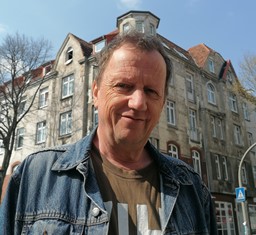 Porträt einer männlich gelesenen Person um die 50, graue kurze Haare, Jeansjacke, T-Shirt. vor einem Altbauhaus.