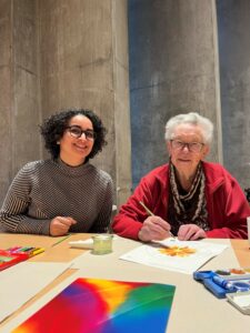 Eine jüngere und eine ältere Frau sitzen nebeneinander an einem Tisch voller Malutensilien und bunter Farben. Die ältere Frau malt mit einem Pinsel.