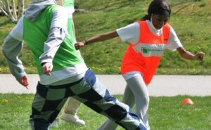 2 Personen, eine älter und in grüner Weste, eine ein Kind in roter Weste, spielen Fußball auf einer grünen Wiese.
