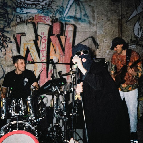 Eine Band vor einer Graffity-Wand. Der Sänger mit Sturmhaube.