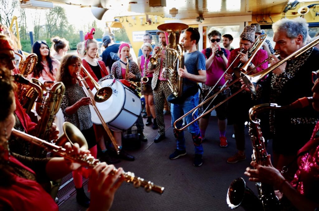 Eine Band, einige mit Animalprint-Klamotten, mit Blechblasinstrumenten, einer Trommel und einer Oboa steht auf dem Deck einer Fähre und macht Musik.