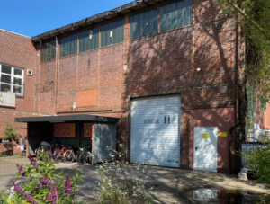 Das Bild zeigt ein altes Industriegebäude aus Backstein mi einem großen weißenTor. In der Mitte einen Fhrradständer, im Vordergrund ein paar Blumen.