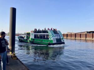 Eine hohe, grün-weiße Fähre mit der Aufschrift "Blankenese" fährt von einem Anleger weg. Auf dem Deck stehen Menschen, die musizieren und Menschen, die zuhören.