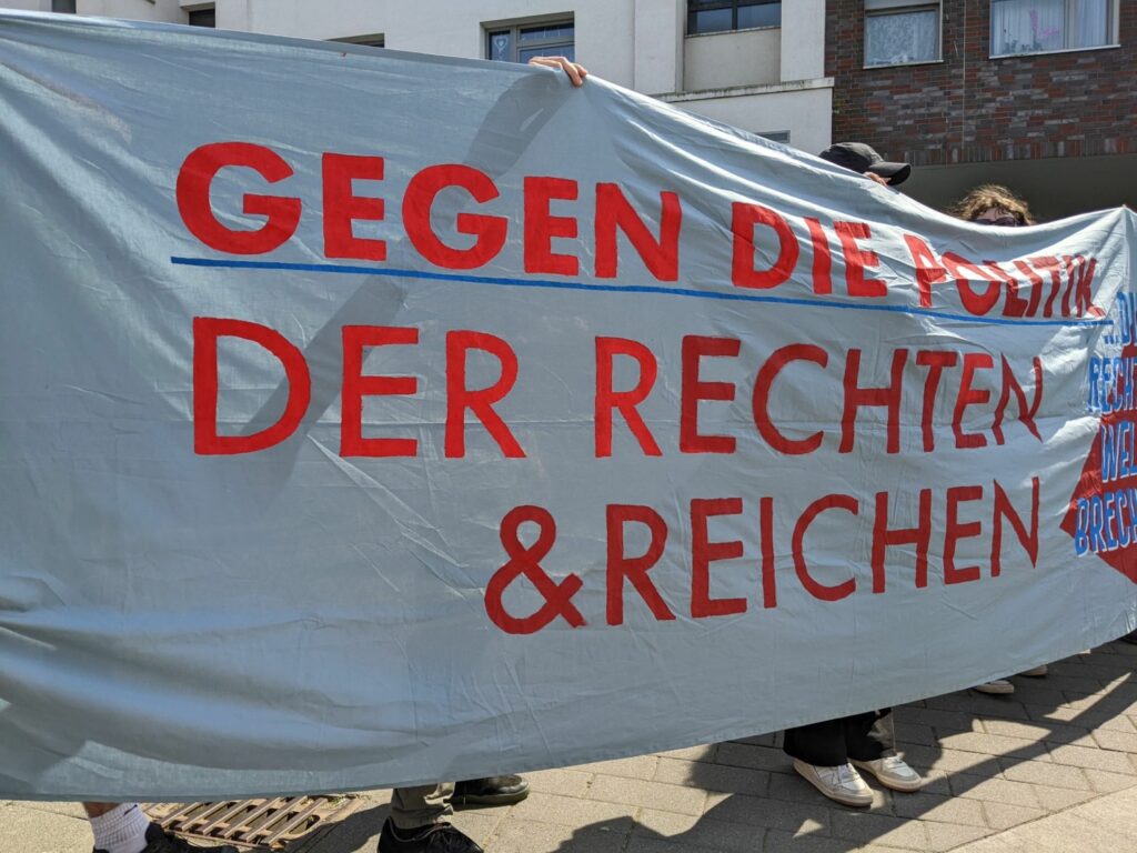 Auf einem großen, hellblauen Transparent steht in roten Großbuchstaben "Gegen die Politik der Rechten und Reichen"