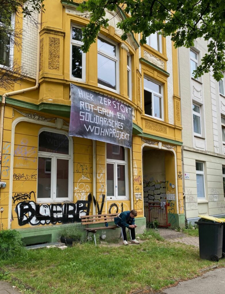 Die Vorderfront an der Fährstraße mit einem Transparent: Hier zerstört Rot-Grün ein solidarisches Wohnprojekt. Auf einer Bank vor dem gelben Gebäude sitzt ein Bewohner mit Handy.