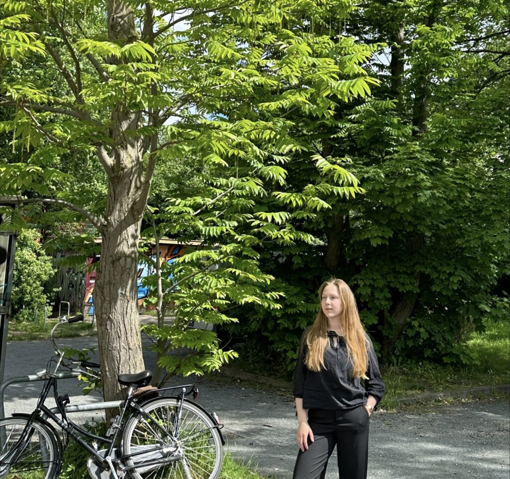 Eine junge Frau, schwarze Hose, schwarze Bluse, lange Haare, steht in einem grünen Park. An einen Baum ist ein schwarzes Fahrrad angelehnt.