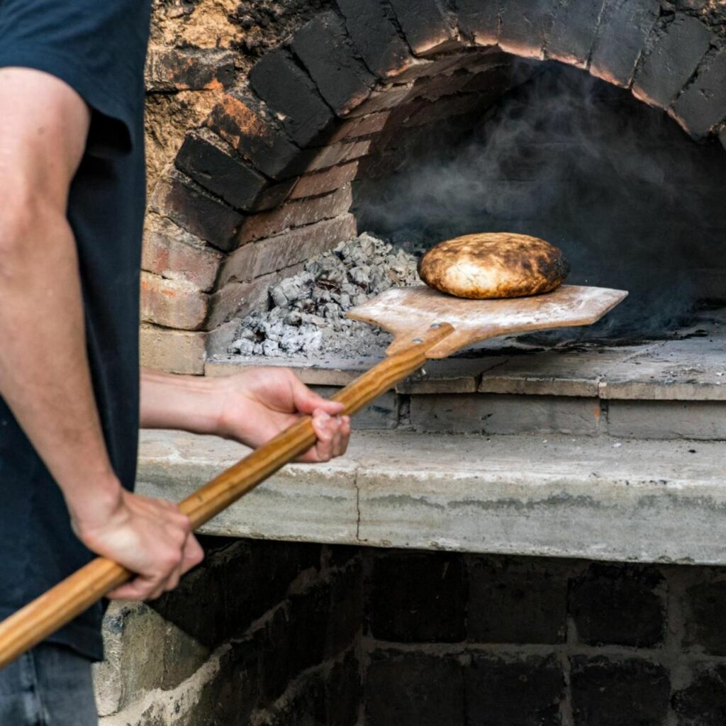 Ein Brot wird mit einem Ofenschieber in einen Holzofen geschoben