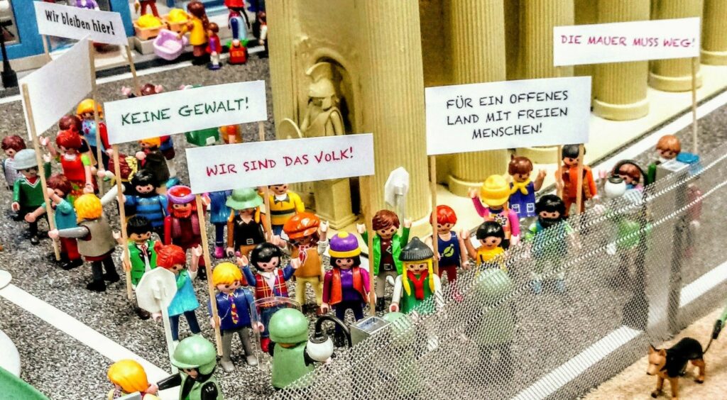 Vor Säulen Playmobilfiguren mit Schildern an Stangen: Keine Gewalt, Wir sind das Volk, für ein offenes Land mit freien Menschen.