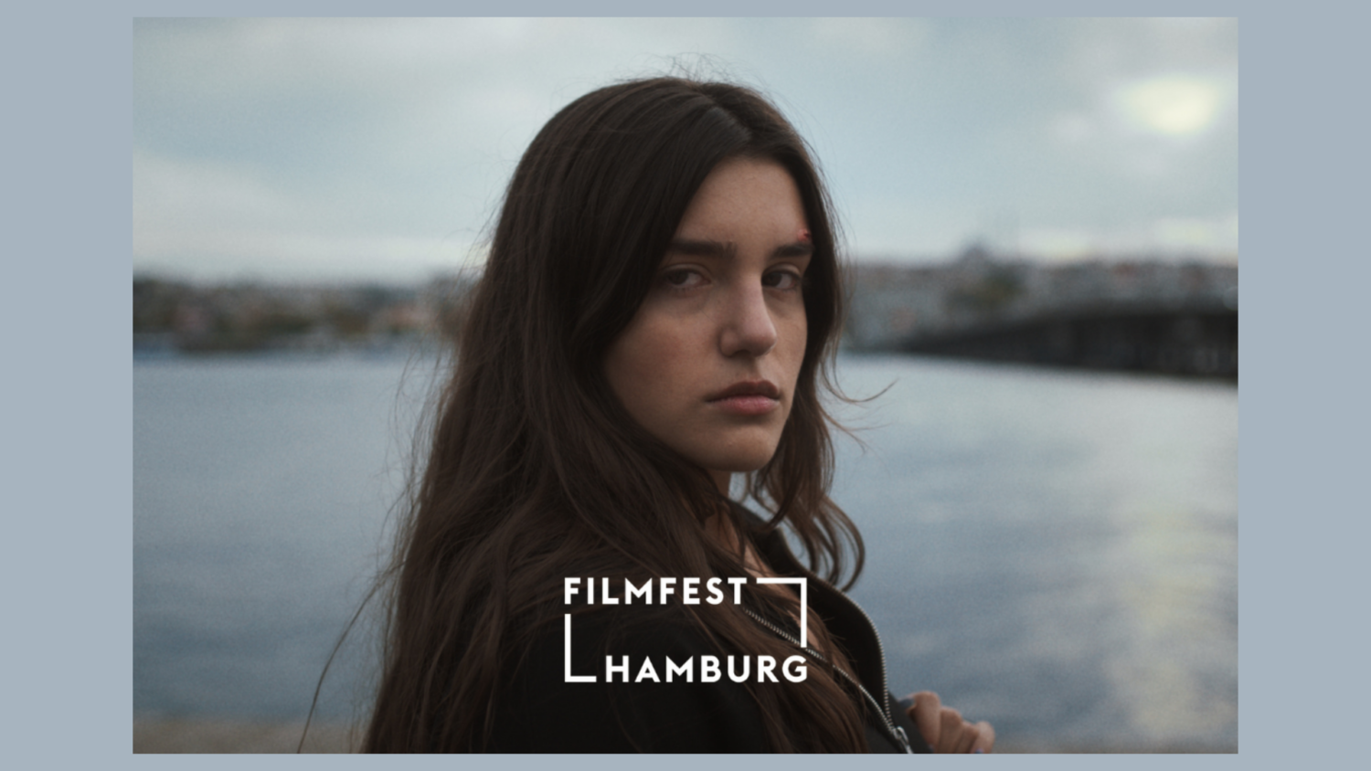 Eine junge Frau, lange braune Haare, skeptischer Blick, blickt über ihre Schulter in die Kamera. Im Hintergrund ein Gewässer, auf das Bild ist der Schriftzug "Filfest Hamburg" aufgebracht.