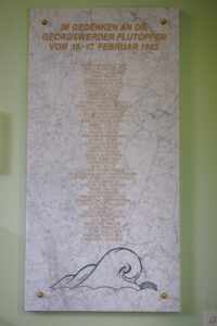 An einer grün gestrichenen Wand ist eine helle Marmortafel geschraubt. Unter der Überschrift: Im Gedenken an die Georgswerder Flutopfer vom 16.-17. Februar 1962, sind alle Namen der Opfer aufgeführt.