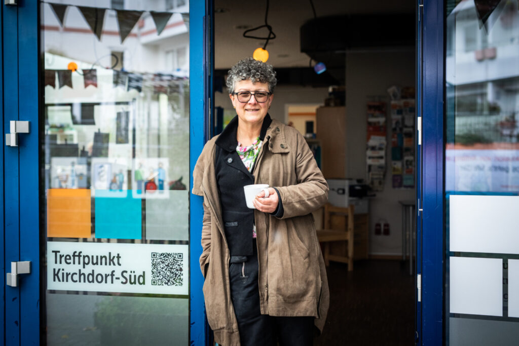 Eine weiblich gelesene Person, lächeln, graue kurze Locken, Brille und beiger Anorak, steht mit einem Becher in der Hand vor einer Glastür mit der Aufschrift "Treffpunkt Kirchdorf-Süd"