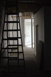 Eine offene Tür wirft Tageslicht in einen dunklen Raum, der nach einer Baustelle aussieht. Eine Leiter ist aufgestellt, Kabel hängen von der Decke