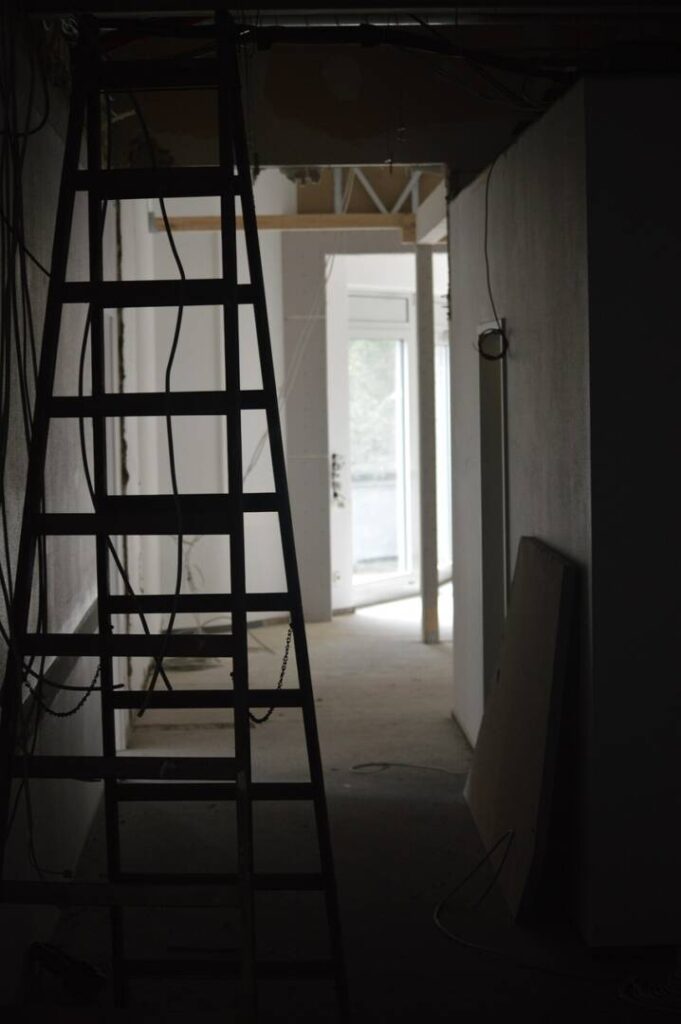 Eine offene Tür wirft Tageslicht in einen dunklen Raum, der nach einer Baustelle aussieht. Eine Leiter ist aufgestellt, Kabel hängen von der Decke
