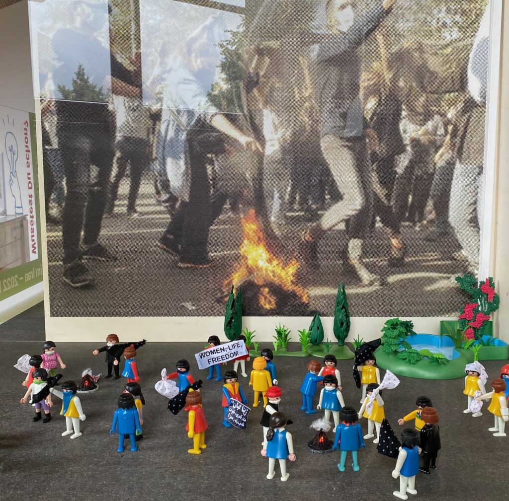 Vor einem großen Bild mit Protestierenden und einem Feuer
wieder Playmobilfiguren.