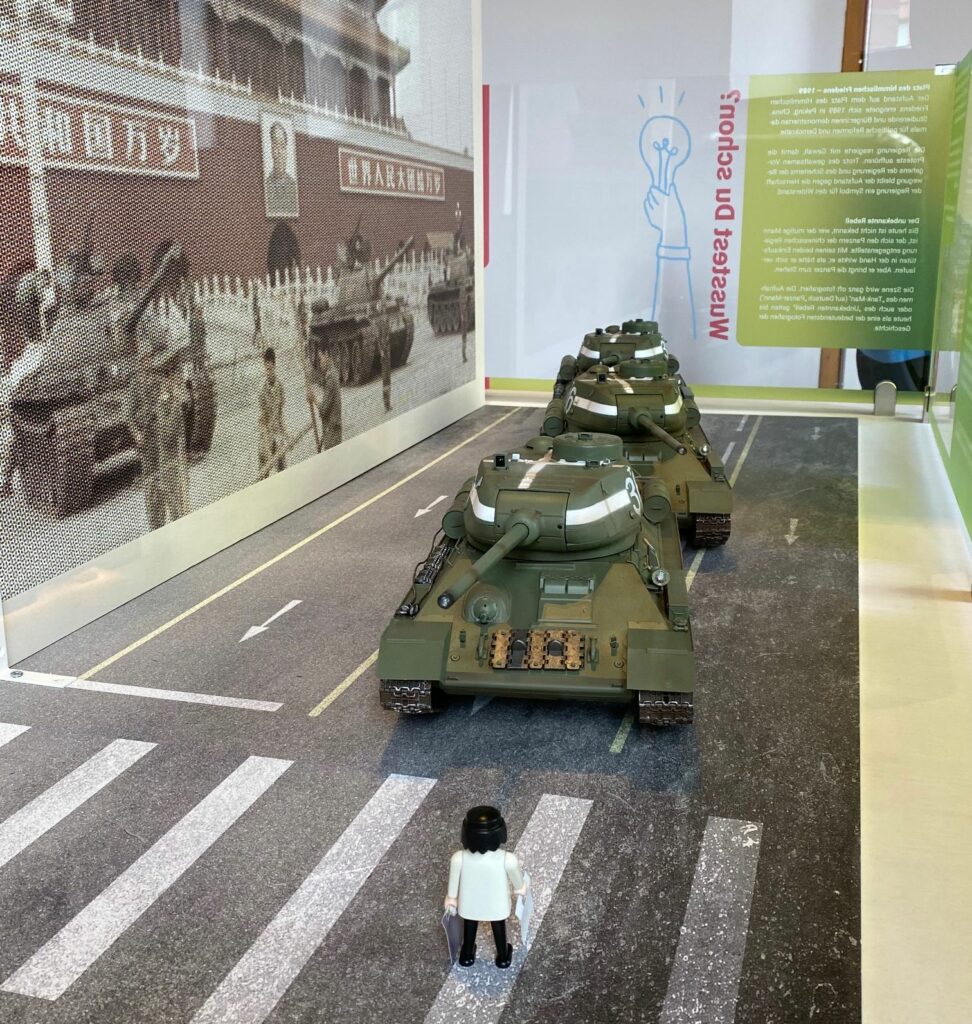 Links ein Gebäude mit chinesischen Schriftzeichen. In der Mitte drei Panzer hintereinander. Davor auf einem Zebrastreifen der Tank Man.