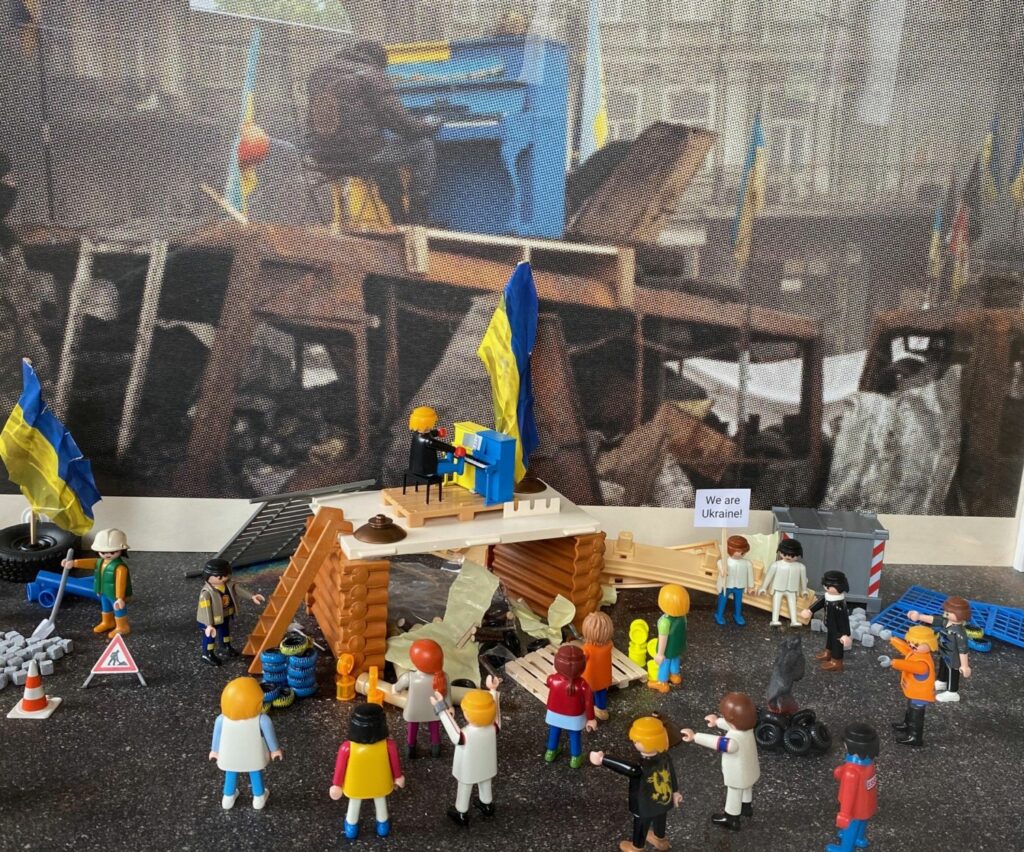 Vorne wieder Playmobilfiguren. In der Mitte ein Gestell, darauf der Klavierspieler mit dem blau-gelben Klavier. Im Hintergrund ein großes Bild mit dem gleichen Motiv.