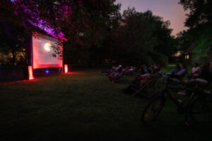 Dunkle Aufnahme aus dem Park, die Leinwand wirft Licht aufs Publikum, das entspannt auf der Wiese in Liegestühlen sitzt 