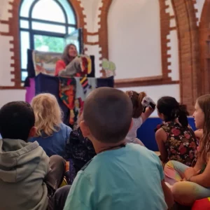 Perspektive aus dem Publikum: Auf der Empore der Kulturkapelle führt eine Frau ein Handpuppentheater vor, Kinder sitzen gespannt und nach vorne gewandt davor