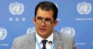 Ein Herr mittleren Alters, dunklekurze Haare, Brille, Anzug, im Sitzen vor ein blauenWand mit den Emblemen der UN