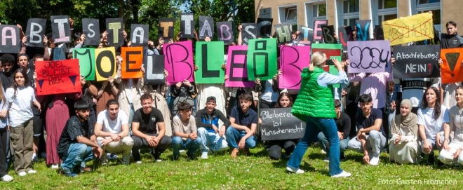 Schüler*innen sitzen und stehen in drei Reihen und halten audff großen bunten Buchstabenschildern „Joel bleibt" hoch.