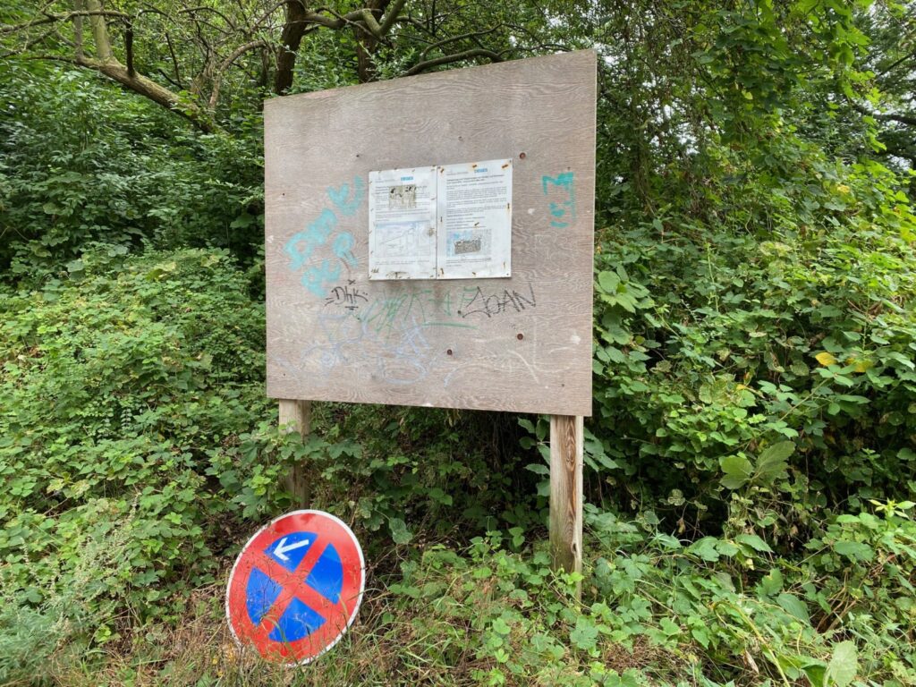 Eine Holz-Plakatwand vor grünen Büschen mit der Baustelleninformation in der Mitte. Darunter auf dem Boden ein Absolutes Halteverbotsschild.