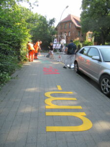 Im Vorgerund in großen gelben Buchstaben auf dem Gehweg die Vorsilbe um- und im Hintergrund in gelb die Silbe ver-.