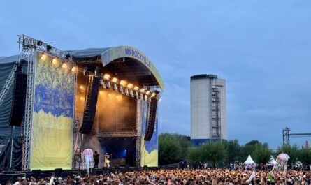 Ein großes Publikum steht vor einer Open-Air-Bühne, es dämmert, im Hintergrund ist ein Getreidesilo und ein Baukran zu sehen.