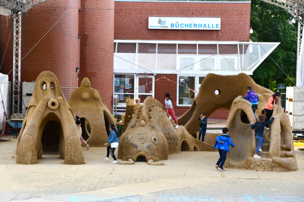 Vor einem Backsteingebäude stehen große Lehmskulpturen, Kinder tummeln sich drum herum.