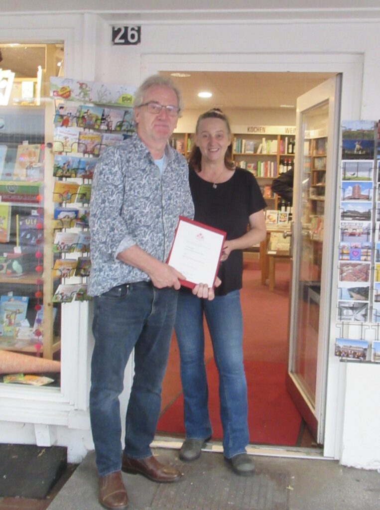 Bede Lüdemann und Teammitglied Sabine Trefzer stehen in der Ladentür. In der Hand halten sie die Urkunde.