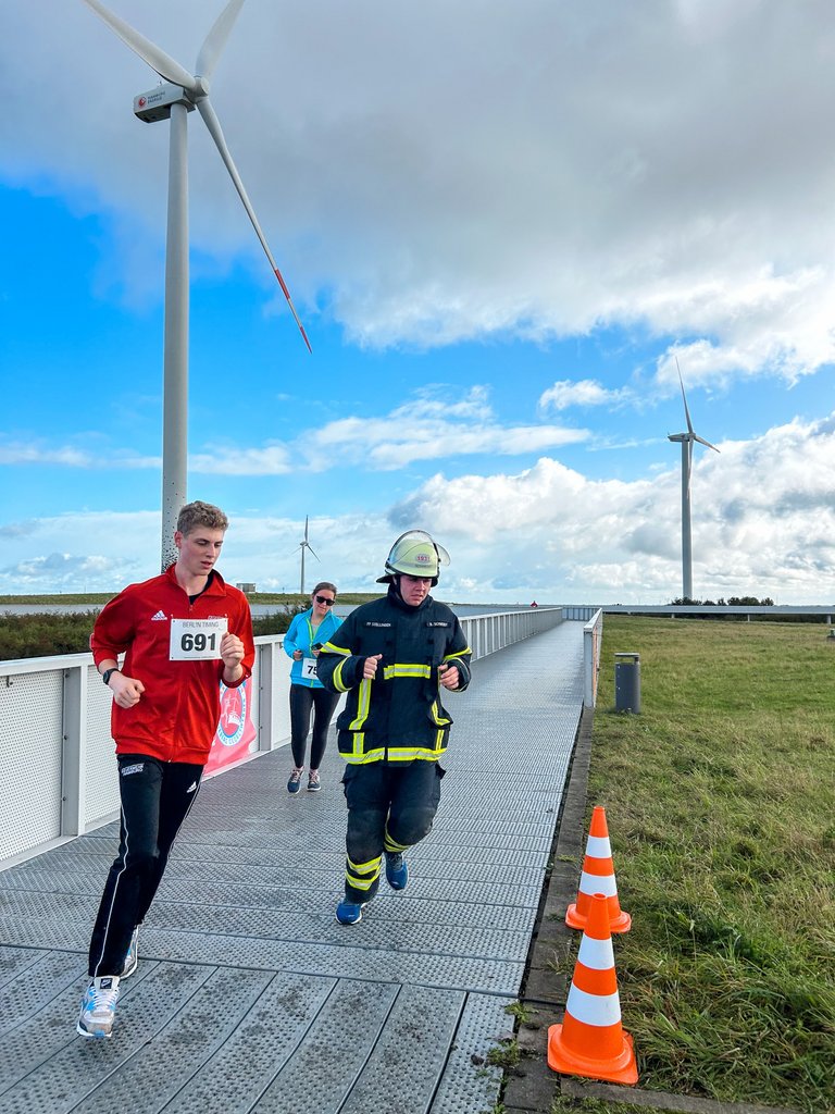 Ein Feuerwehrmann und eine Person im Laufoutfit laufen auf einer geraden Strecke, die von Wiese umgeben ist. Blauer Himmel mit weißen Wolken