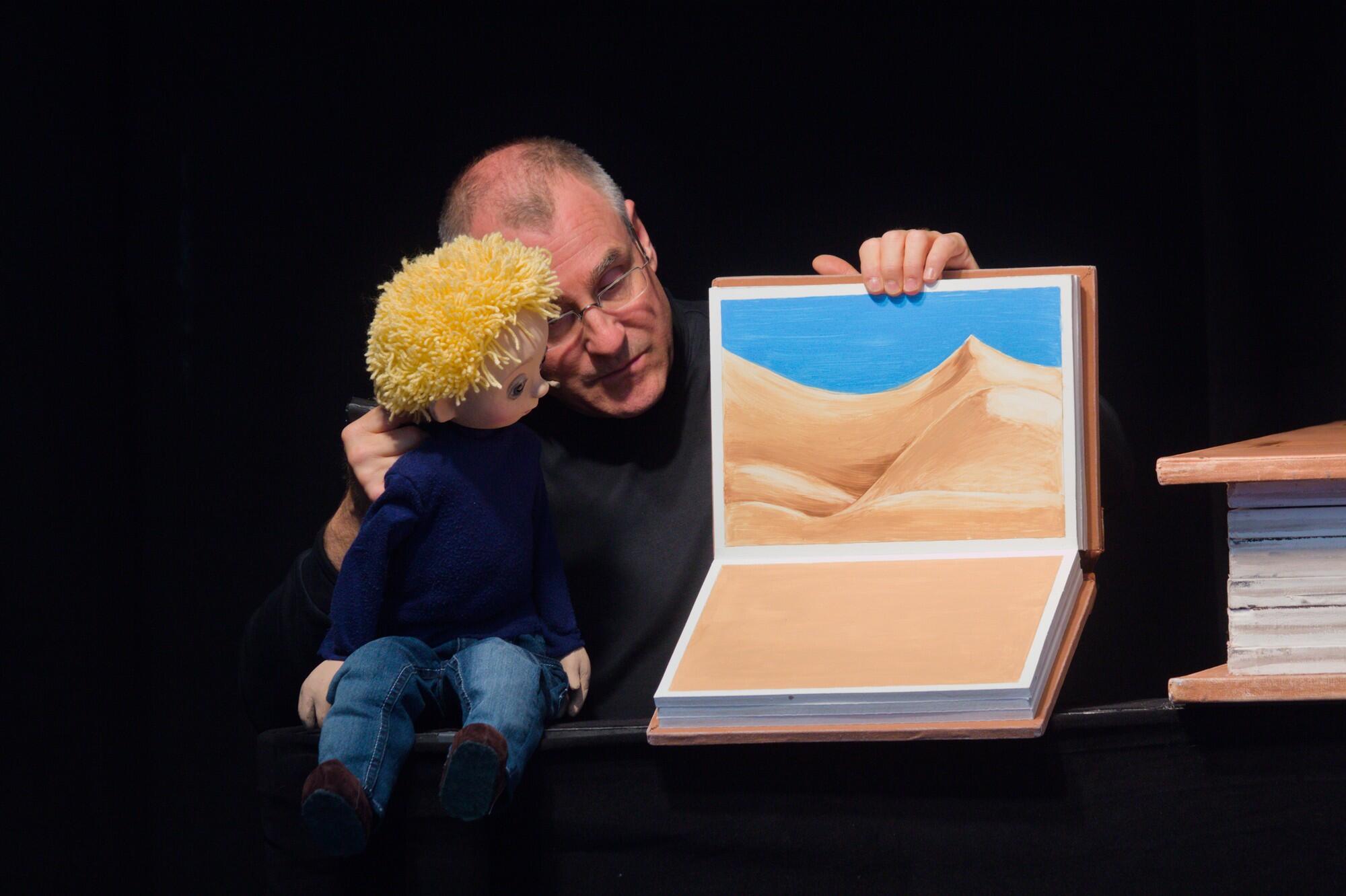 Ein Mann mit einer Handpuppe, der ein kleines Kind ist, hält ein Buch offen hoch, in dem eine Seite mit Wüste und blauem Himmel abgebildet ist.