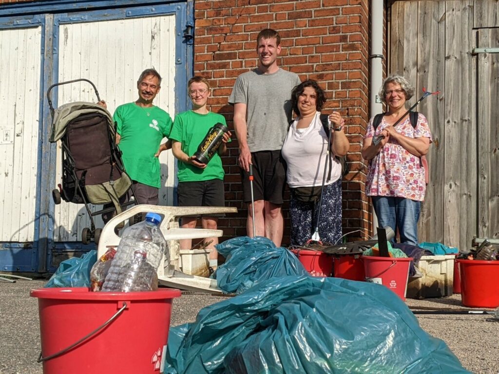 Von Menschen stehen vor einem Ziegelbau mit großen Holztüren (ein Ruderverein), einer von ihnen hält einen Kinderwagen in die Luft, eine andere hält eine Gaskartusche in den Händen. Ein Paar hält sich im Arm, eine Frau hat eine Greifzange über der Schulter. Vor ihnen liegt ein Plastikstuhl, mehrere blaue gefüllte Müllsäcke und rote Eimer aus denen Müll ragt.