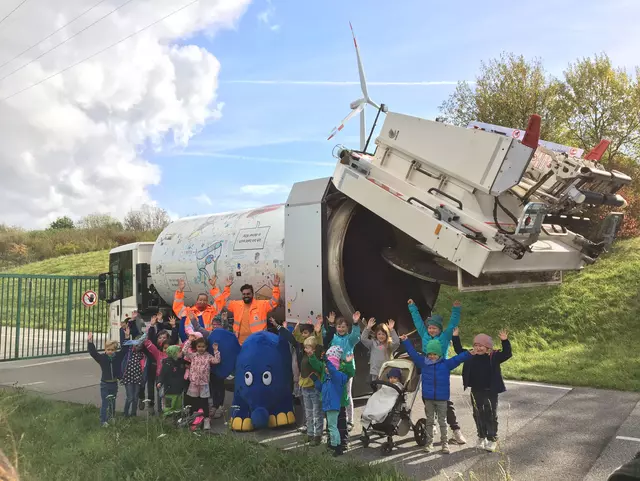 Vor einem Müllfahrzeug stehen 2 Menschen in orangener Kleidung, viele Kinder und ein großer blauer Stoffelefant