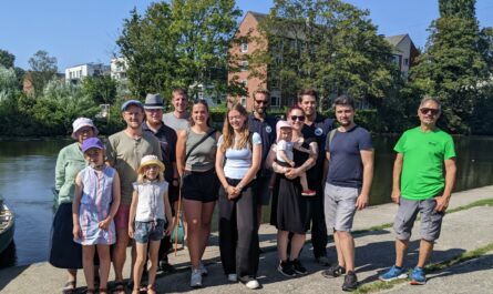 Eine Gruppe Menschen, Kinder und Erwachsene jeden Alters, stehen in sommerlicher Kleidung auf einem Betonablass, dahinter ein Kanal , im Hintergrund ein Mehrfamilienhaus und grüne Bäume. Der Himmel ist blau, die Sonne scheint.