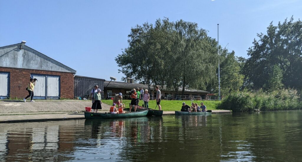 Bild vom Wasser aus: Mehrere Menschen in Rettungswesten steigen in Kajaks, währens sie ein Kamerateam filmt