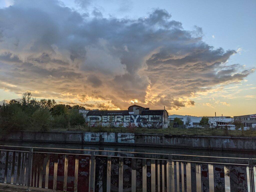 Über einer alten, mit Graffiti besprühten Lagerhalle auf der anderen Seite eines Kanals ziehen sich Wolken am abendlichen Himmel zusammen