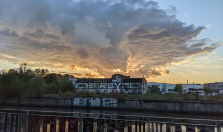 Über einer alten, mit Graffiti besprühten Lagerhalle auf der anderen Seite eines Kanals ziehen sich Wolken am abendlichen Himmel zusammen