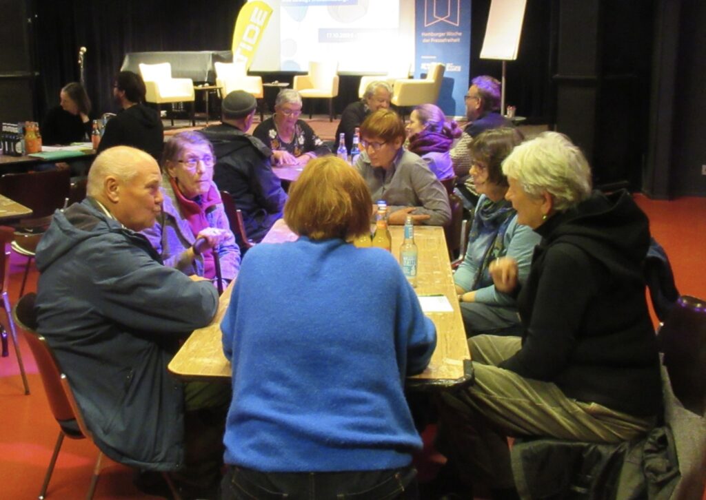 Im Vordergrund eine Diskussionsrunde mit sechs Beteiligten an einem länglichen Tisch. Im Hintergrund weitere Tischrunden