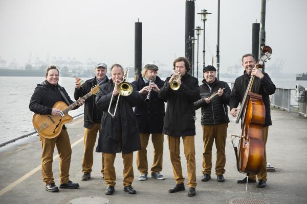 Sieben Männer mit schwarzen Oberteilen und braunen Hosen stehen am Hafen mit ihren Instrumenten: Kontrabass, Trompete, Gitarre, Klarinette, Posaune. Ein grauer Tag, im Hintergrund Conatinerbrücken, um den Steg herum Wasser. Die Männer haben gute Laune.