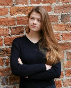 Portait einer jungen Frau, schwarze Kleidung, lange Haare, vor einer roten Backsteinwand