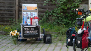 Ein Bollerwagen, auf dem "Mobiler Erinnerungsort, Außenszelle Dessauer Ufer" steht. Darauf Isokanne, Hafermilch und Infobroschüren, dabenem ein Eimer mit Blumen und eine Kiste mit Selterflaschen.